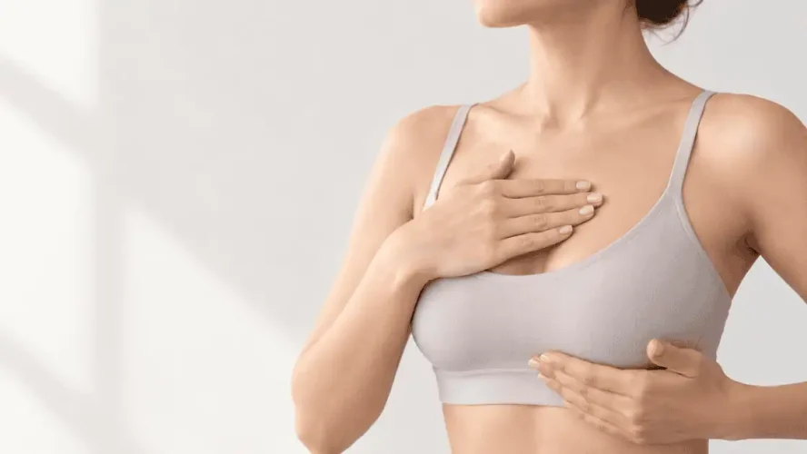 Woman in a neutral top gently touching her chest during breast augmentation recovery